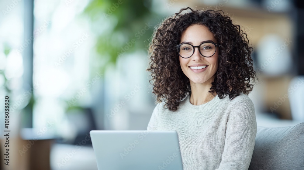 Cheerful woman with curly hair working on a laptop in a modern workspace, bright and inviting atmosphere.