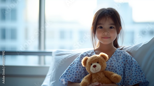 A young Asian girl sits comfortably in a hospital bed, holding a teddy bear closely. Sunlight filters through the window, creating a warm and hopeful atmosphere as she rests photo