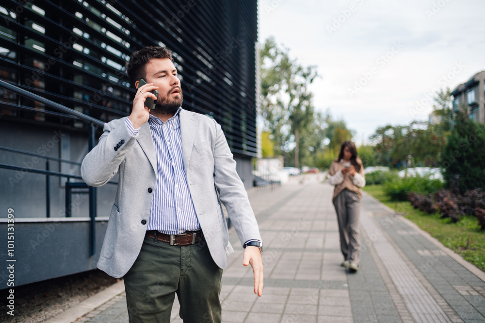 Businessman having annoyed phone call outside office with woman walking away