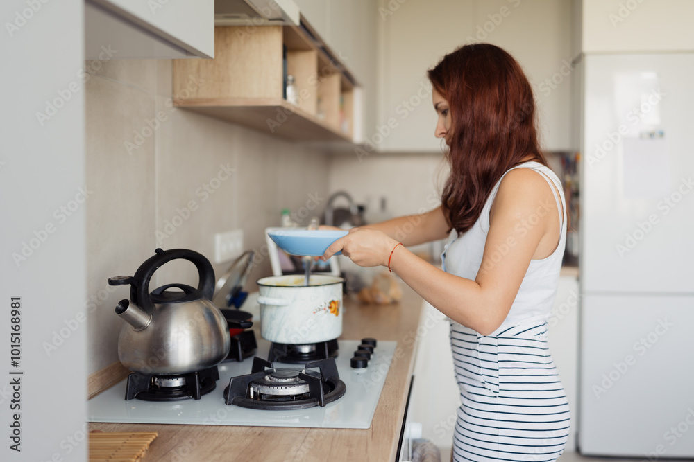A woman with red hair is preparing food on a stove in a modern kitchen. She is holding a bowl while standing near a kettle and pot A woman with red hair is preparing food on a stove in a modern kitchen. She is holding a bowl while standing near a kettle and pot