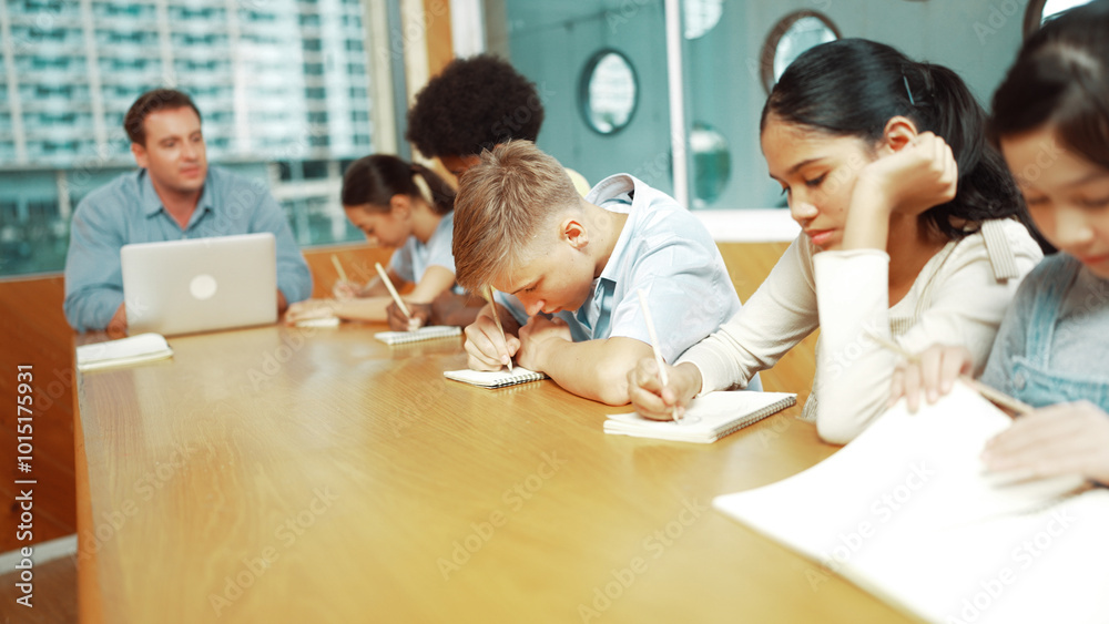 Young smart teenager writing and talking about topic at classroom. Diverse highschool student taking a note in other notebook while sitting with multicultural friends. Creative education. Edification. Young smart teenager writing and talking about topic at classroom. Diverse highschool student taking a note in other notebook while sitting with multicultural friends. Creative education. Edification.
