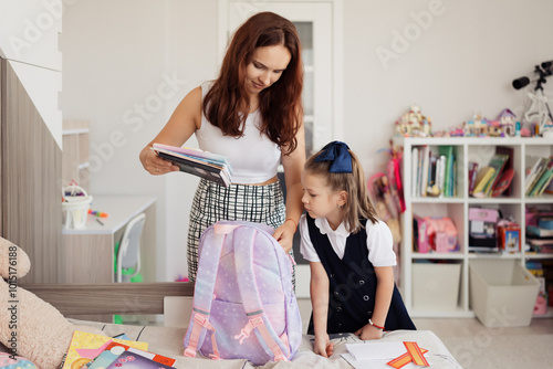 A mother assists her daughter in packing a backpack in preparation for school. The room is organized, with colorful books and school supplies visible, symbolizing learning and preparation