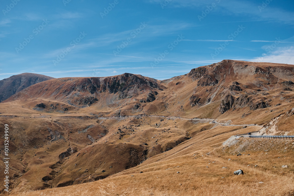 Stunning panoramic view of a winding road through a mountain pass. Rugged peaks, dry grass, and a clear blue sky create a picturesque scene. Parâng Mountains, Romania. Transalpina road. Stunning panoramic view of a winding road through a mountain pass. Rugged peaks, dry grass, and a clear blue sky create a picturesque scene. Parâng Mountains, Romania. Transalpina road.