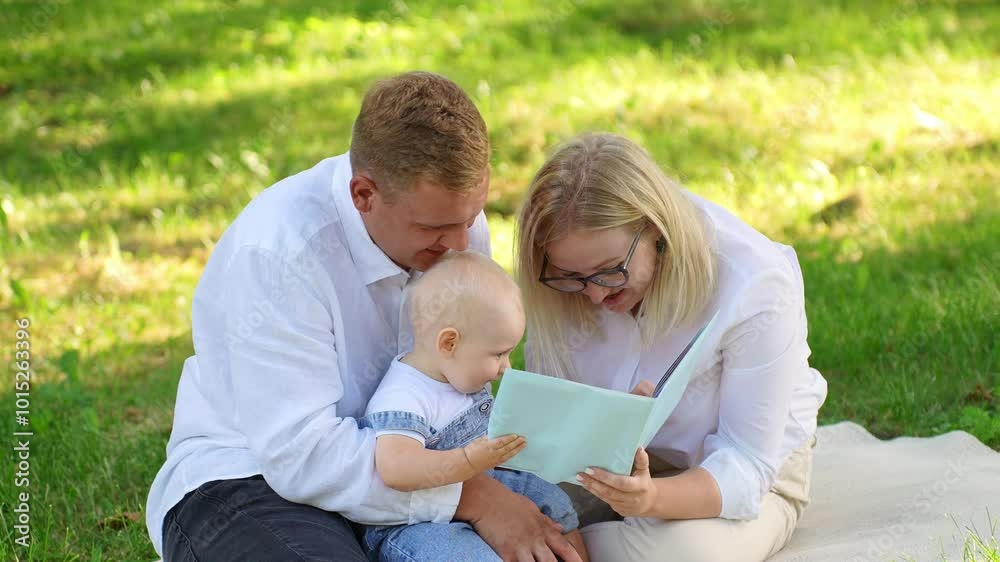 Parents reading book to their child while sitting on blanket during picnic in park, watching educational pictures, close-up. Weekend in nature. Happy family concept, infant education. Parents reading book to their child while sitting on blanket during picnic in park, watching educational pictures, close-up. Weekend in nature. Happy family concept, infant education.