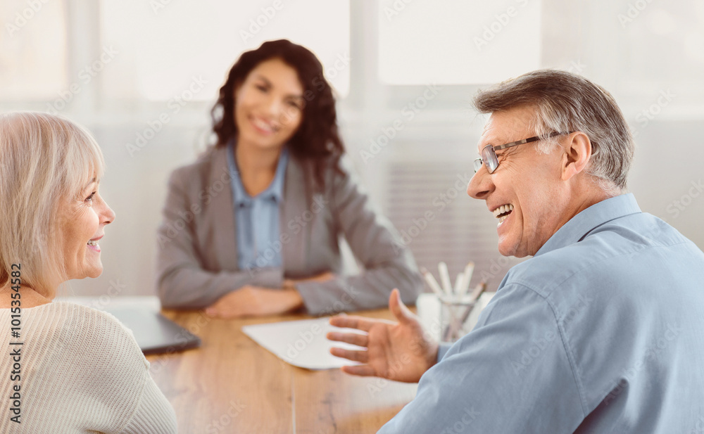 Good News. Happy senior couple looking at each other and listening to saleswoman sitting at the desk in the background Good News. Happy senior couple looking at each other and listening to saleswoman sitting at the desk in the background