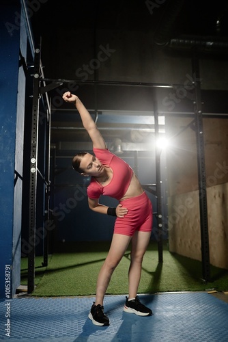 Vertical shot of young sporty woman wearing pink crop top and matching shorts warming up side bending while stretching muscles for daily training on bars in gym, copy space