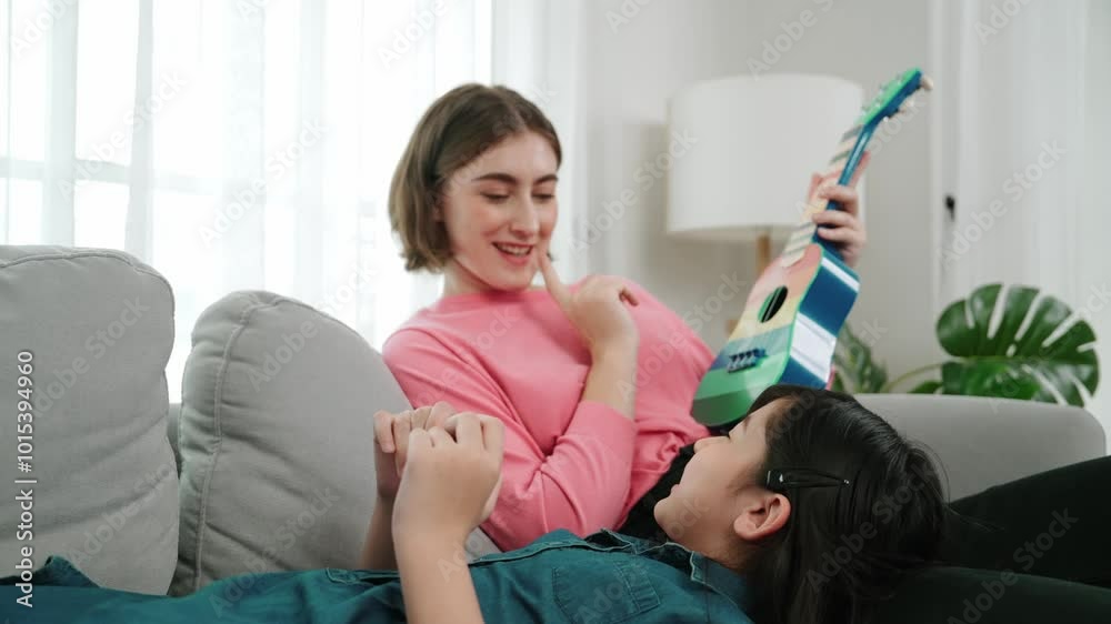 Asian cute daughter listening acoustic music while lie on her mother lap at sofa. Caucasian mother playing colorful ukulele while cute child looking at her parent. Family recreation concept. Pedagogy.