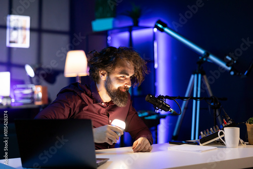 Content creator with freakish ruffled hair holding smart light bulb under face to create creepy mood in studio while telling horror story. Disheveled man in apartment using light to create spooky vibe