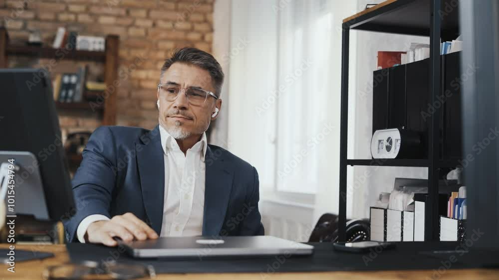 Smiling businessman wearing suit and glasses working on laptop in loft office. Corporate manager focusing on work. Happy mid adult man sitting at desk, smiling. Male entrepreneur managing business. Smiling businessman wearing suit and glasses working on laptop in loft office. Corporate manager focusing on work. Happy mid adult man sitting at desk, smiling. Male entrepreneur managing business.
