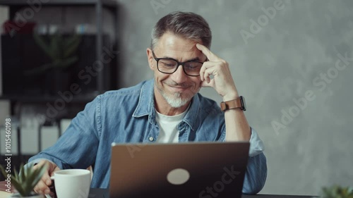Portrait of mature man with grey hair and beard smiling. Confident businessman working on laptop from home office. Male entrepreneur sitting at desk in bright room wearing casual denim shirt.