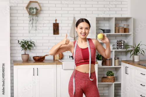 Young woman in sportswear, with apple and measuring tape in kitchen. Weight loss concept