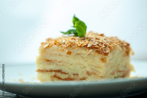 Multi layered napoleon cake with a crispy crust and delicate custard is being decorated with a sprig of mint on a white plate