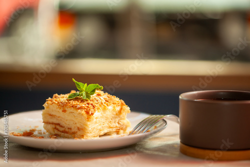 Indulge in a decadent napoleon cake with mint on a wooden plate next to tea in a cozy cafe. Creamy custard, flaky pastry, and refreshing mint make the perfect bite. Enjoy a sweet moment