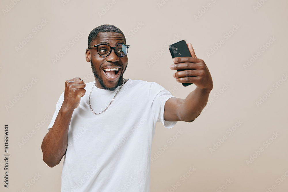 Excited young man taking a selfie, smiling joyfully, wearing glasses and a casual white t shirt, showing enthusiasm and happiness against a neutral beige background Excited young man taking a selfie, smiling joyfully, wearing glasses and a casual white t shirt, showing enthusiasm and happiness against a neutral beige background