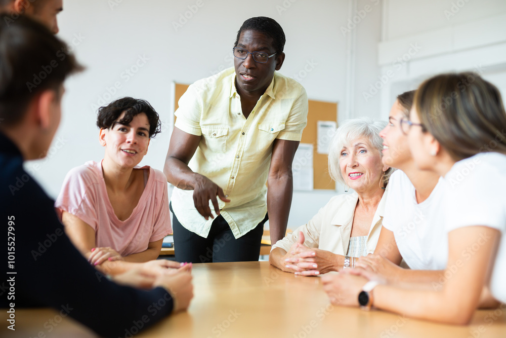 Fototapeta premium Positive friendly African American English tutor giving language courses to group of interested adults of different ages and nationalities sitting around table in classroom..