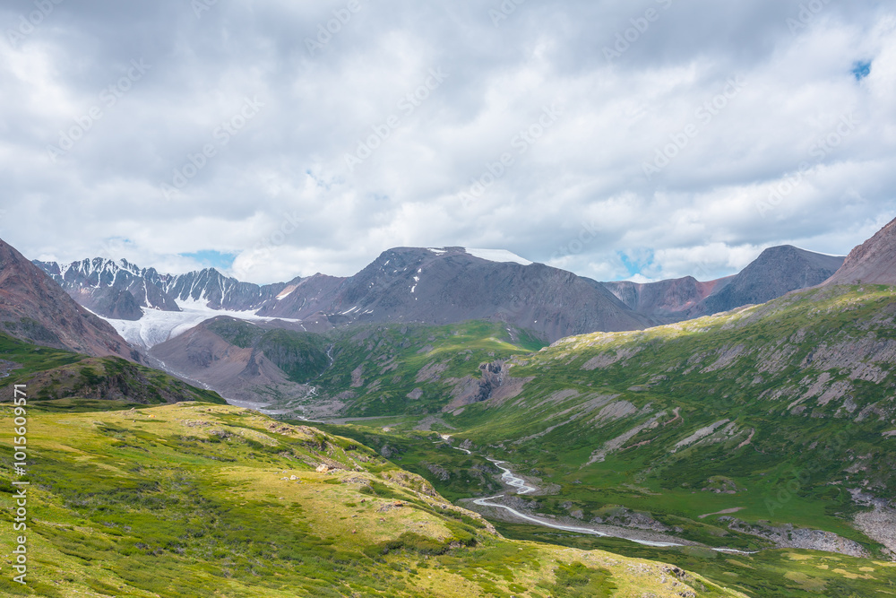 Fototapeta premium Wide green alpine valley with snake river among hills and rocks with vast view to large snow mountain top and big glacier tongue under cloudy sky. Serpentine river in high mountains. Shadows of clouds