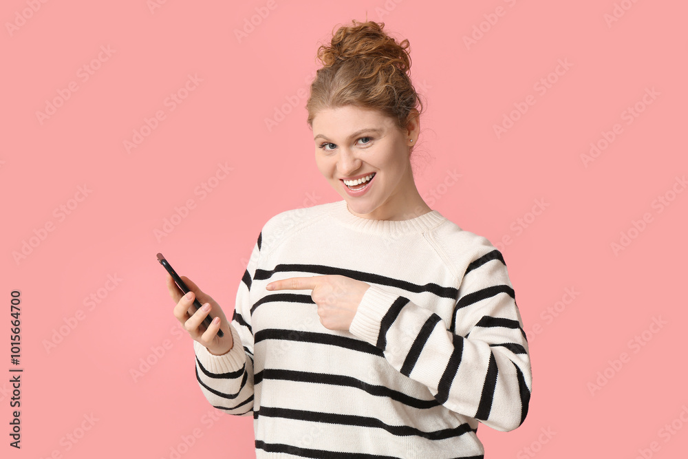 Happy beautiful young woman pointing at mobile phone on pink background