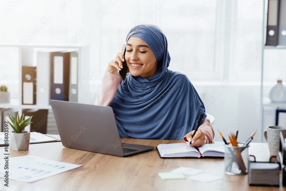 Business Lifestyle. Portrait Of Busy Middle Eastern Businesswoman At Workplace In Office, Talking On Cellphone, Using Laptop And Taking Notes Business Lifestyle. Portrait Of Busy Middle Eastern Businesswoman At Workplace In Office, Talking On Cellphone, Using Laptop And Taking Notes
