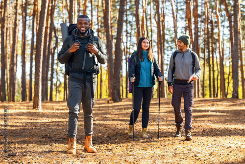 Cheerful multiethnic backpackers walking by autumn forest, full length photo, active lifestyle