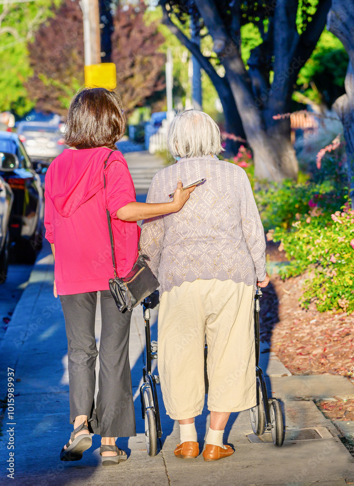 Senior woman walking using a mobility walker on the pedestrian footpath, chatting to her daughter. California. Vertical format.