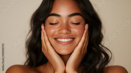 Joyful young woman with glowing skin and dark wavy hair smiling softly, hands resting on her face