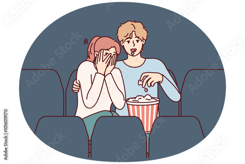 Guy and girl are watching horror movie in cinema and eating popcorn enjoying films about vampires. Fearful woman closes eyes with hands sitting in cinema near man watching horror movie with interest