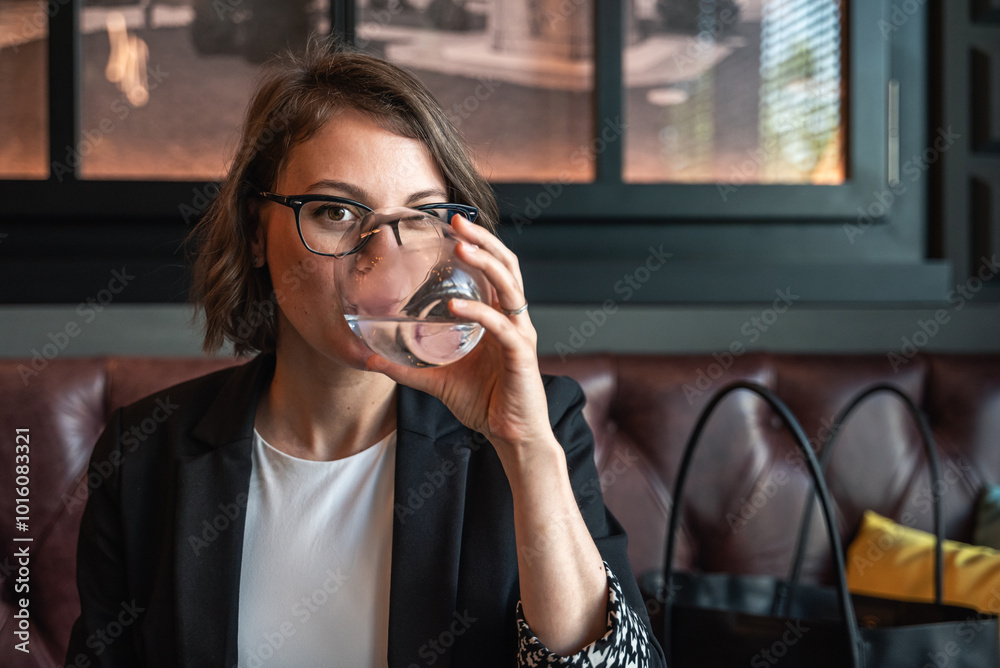Woman, drinking water in office or a cafe, thinking for hydration, wellness or finance company. Accountant holding a glass of water and looking at the camera. Woman, drinking water in office or a cafe, thinking for hydration, wellness or finance company. Accountant holding a glass of water and looking at the camera.
