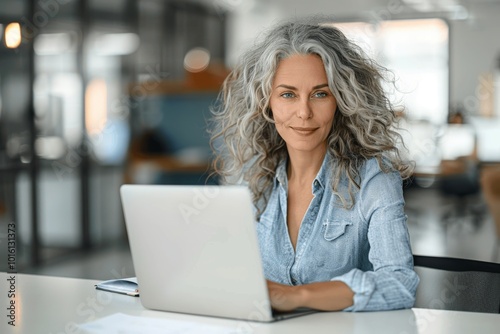 Joyful Middle-Aged Woman with Grey Curly Hair Working on Laptop at White Desk in Modern Light Office