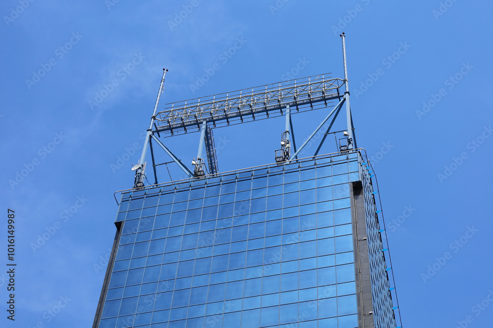 The top of high modern skyscraper made of blue toned glass windows and steel constructions under blue sky. Perspective view from below to office building roof with antennas.