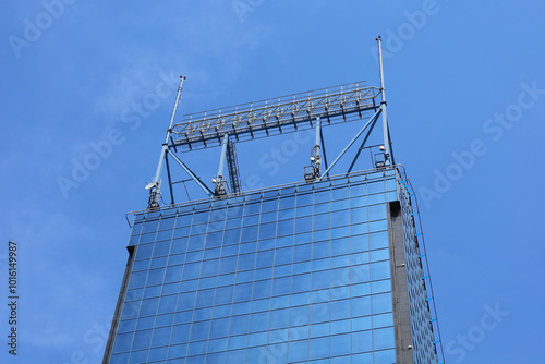 Obraz na płótnie The top of high modern skyscraper made of blue toned glass windows and steel constructions under blue sky. Perspective view from below to office building roof with antennas. w salonie