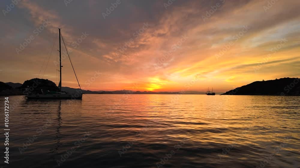 Turkiye, Marmaris, Sunrise at Kadirga Bay, a sailboat and the land silhouettes during sunrise, a sailboat is leaving the bay slowly,so tranquil and peaceful. Colors from blue to yellow to orange. Turkiye, Marmaris, Sunrise at Kadirga Bay, a sailboat and the land silhouettes during sunrise, a sailboat is leaving the bay slowly,so tranquil and peaceful. Colors from blue to yellow to orange.