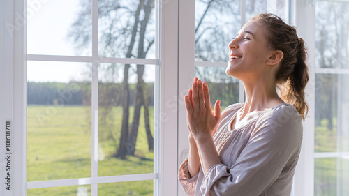 woman with light-colored clothes standing in front of a sunny window, with her hands clasped together praying