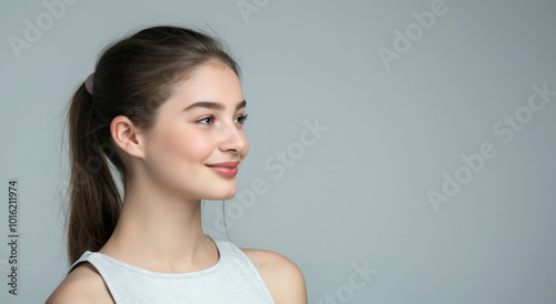 Side profile of a smiling young woman with long brown hair tied in a ponytail, wearing a white tank top. The neutral background and soft lighting emphasize her natural beauty and calm expression.