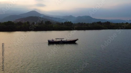 Wallpaper Mural A long fishing boat glides smoothly up the river as the sun sets behind Bokor Mountain. The sky is filled with clouds, reflecting vibrant colors over the water, creating a serene atmosphere. Torontodigital.ca