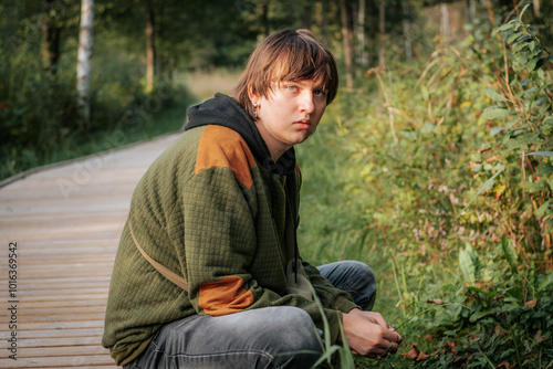 A teenage boy is sitting on the edge of a wooden path surrounded by greenery, wearing a green and orange jacket. He gazes thoughtfully into the distance, immersed in the natural environment.