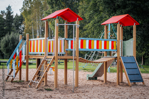 A lively playground with colorful slides, ladders and climbing equipment. The structure has red roofs and bright panels set on a sandy surface with trees in the background.