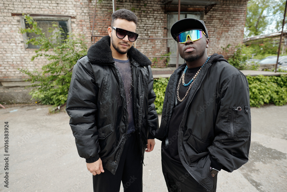 Portrait of two men standing outside an old brick building wearing fashionable jackets and sunglasses sharing a confident pose without smiling Portrait of two men standing outside an old brick building wearing fashionable jackets and sunglasses sharing a confident pose without smiling