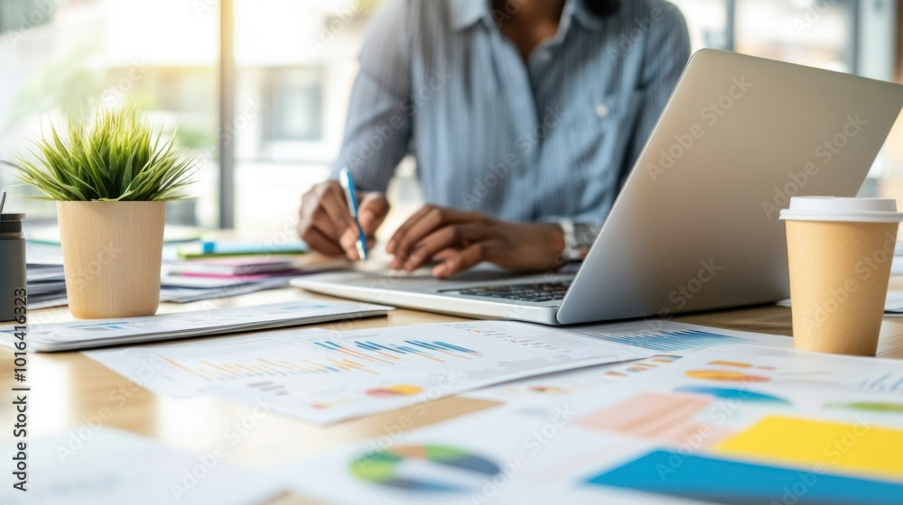 A person working on a laptop at a desk surrounded by charts, plants, and coffee cups. A person working on a laptop at a desk surrounded by charts, plants, and coffee cups.
