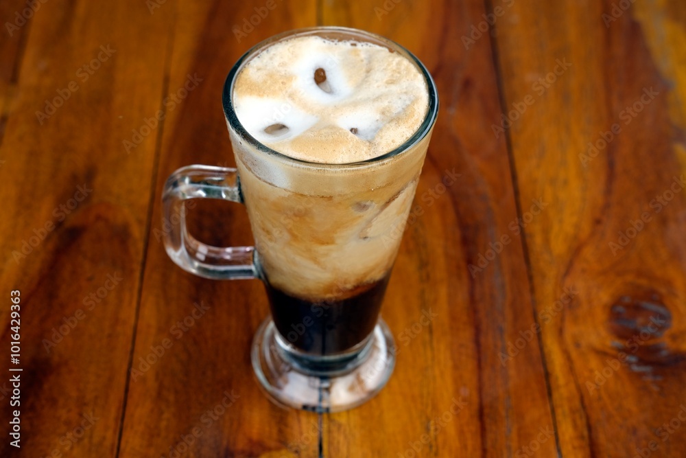A refreshing glass of iced coffee with creamy layers, served on a wooden table. The lush green background adds a natural, serene atmosphere, perfect for enjoying a cold brew on a sunny day. A refreshing glass of iced coffee with creamy layers, served on a wooden table. The lush green background adds a natural, serene atmosphere, perfect for enjoying a cold brew on a sunny day.