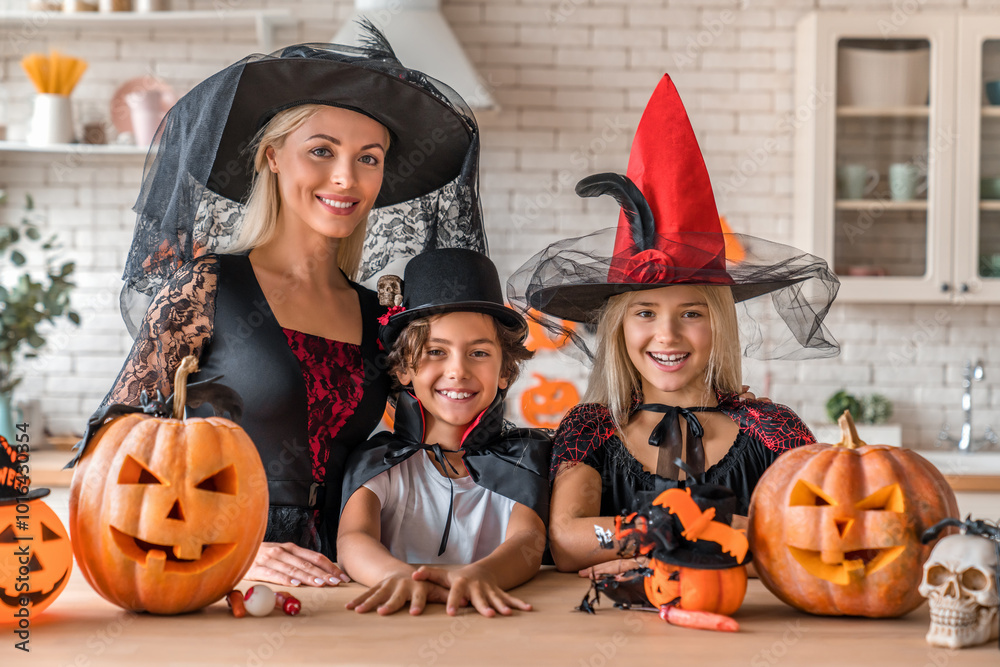 Fototapeta premium Adult mother and children standing in witch vampire costumes at the kitchen and looking in camera. Festive Halloween celebration with costumed family and pumpkin decorations in cozy kitchen