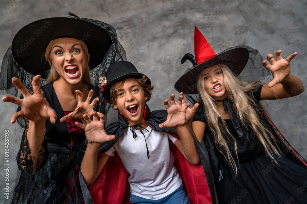 Obraz premium Happy family having fun scaring with look in camera standing over grey background. Mother and children kids in witch vampire costumes and makeup on a celebration of Halloween on dark grey background