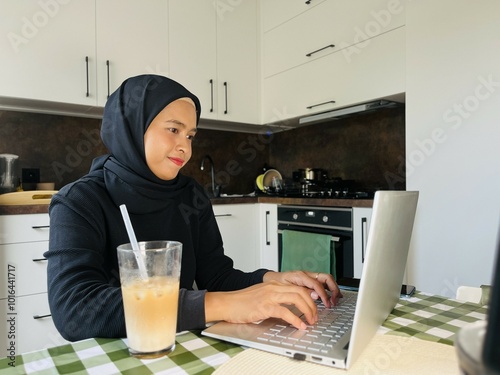 Happy Asian woman while working at home. Young woman using laptop in the kitchen. Typing on the keyboard laptop. Iced coffee beside the laptop.  photo