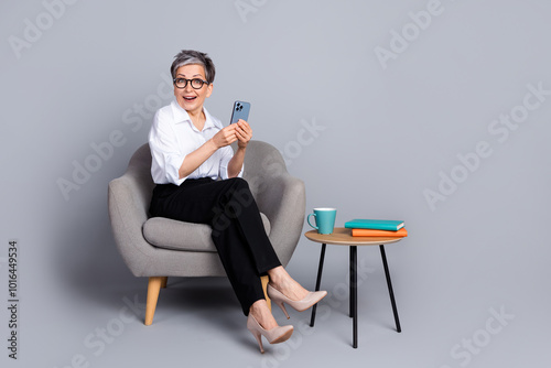 Full length photo of lovely senior lady sit armchair hold device amazed dressed stylish formalwear isolated on gray color background photo