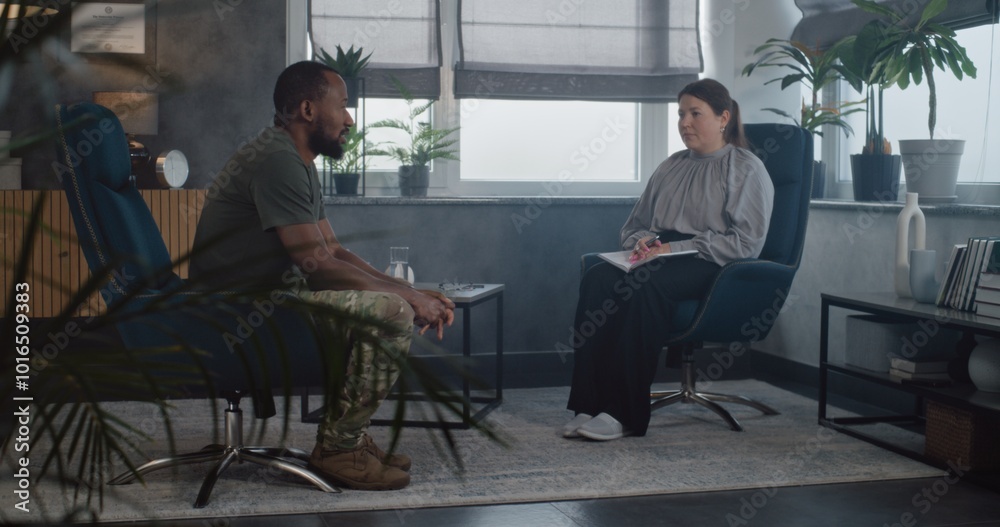 Female psychologist sits in chair with notebook, provides counseling session, listens military man. African American male soldier with PTSD talks to psychotherapist during therapy. Psychological help. Female psychologist sits in chair with notebook, provides counseling session, listens military man. African American male soldier with PTSD talks to psychotherapist during therapy. Psychological help.