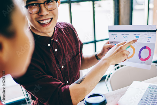 Cheerful asian hipster guy with paper diagram in hand laughing during cooperation with female partner, successful financial experts discussing data raport during accounting process near desktop photo