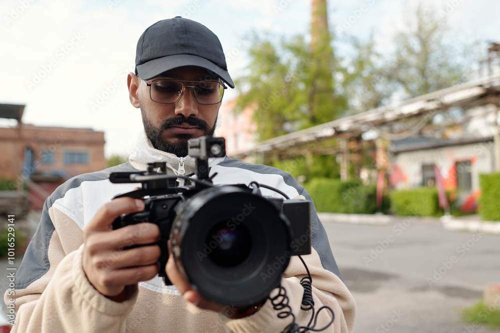 Man wearing hat and glasses holding professional camera while standing outdoors, adjusting settings with focused expression, blurry background of buildings and trees adding depth Man wearing hat and glasses holding professional camera while standing outdoors, adjusting settings with focused expression, blurry background of buildings and trees adding depth