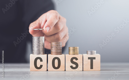 A hand carefully stacking coins on a wooden block labeled 'COST,' representing the concept of financial management, budgeting, and the importance of controlling expenses photo