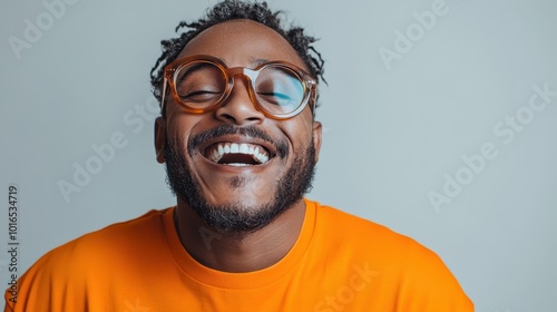 A man with a trimmed beard and round glasses smiles broadly in a portrait, wearing an orange shirt, exuding friendliness and warmth against a neutral background.