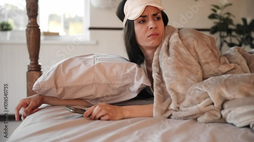 A woman lies in bed under a blanket, wearing a sleep mask on her head, and looking frustrated at her phone. The scene suggests early morning, possibly waking up to an unwelcome notification or alarm.