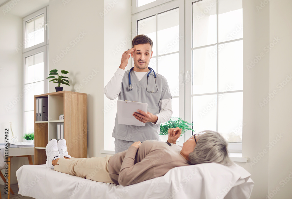 Senior elderly gray-haired woman patient lying on couch in medical office talking to a male doctor holding report file with appointment and giving consultation during medical examination in clinic. Senior elderly gray-haired woman patient lying on couch in medical office talking to a male doctor holding report file with appointment and giving consultation during medical examination in clinic.
